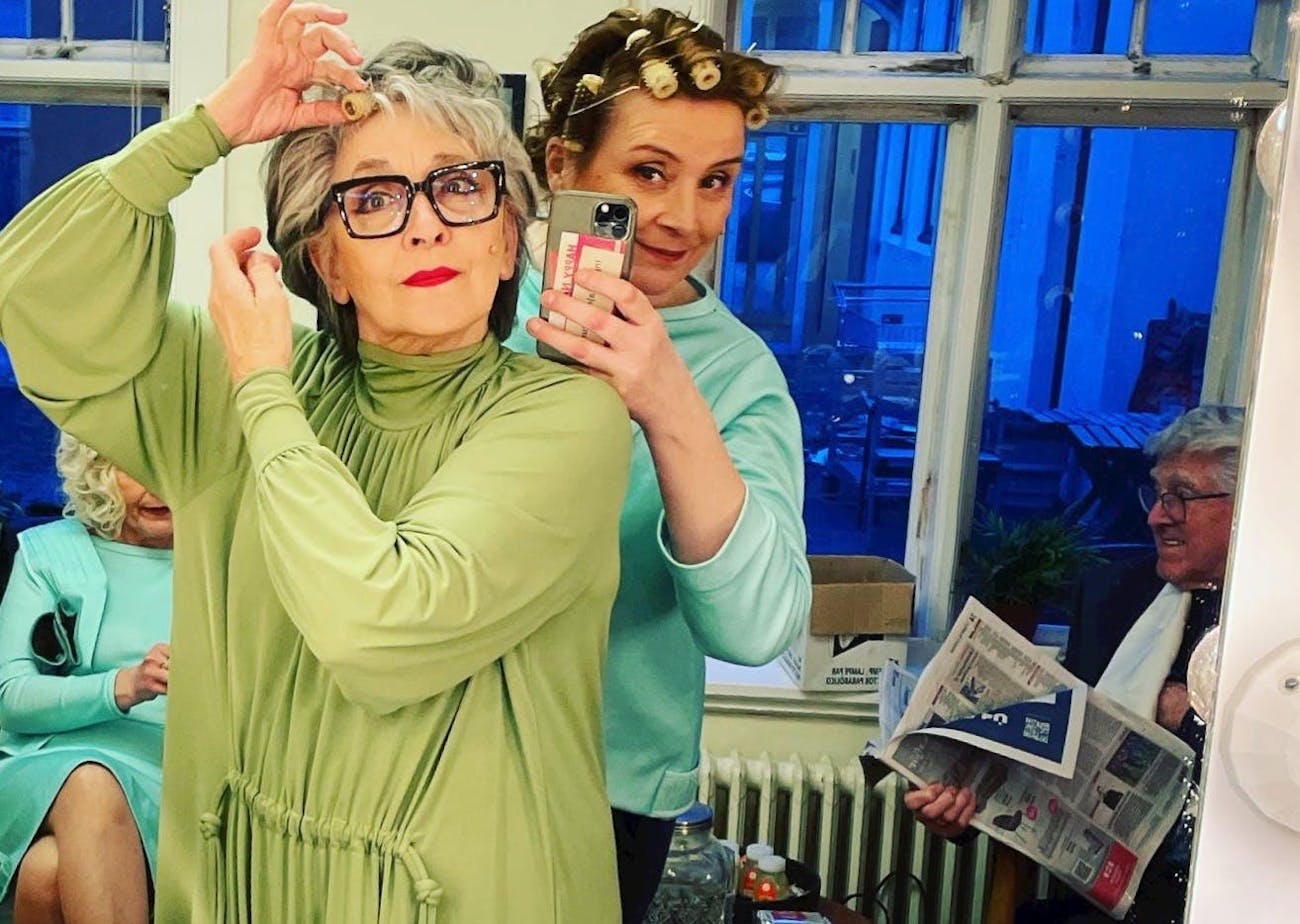 Picture of two persons in the foreground. They are looking at each other in a mirror. The person in front wears green dress and glasses and red lipstick. The person takes a hair roll from its grey hair. The second person right behind is wearing a Turkish blue sweater and had hair rolls in brown hair. That persons holds a telephone and take the picture. In the background one can see two persons sitting. One in a Turkish blue dress and another one holding a news paper.