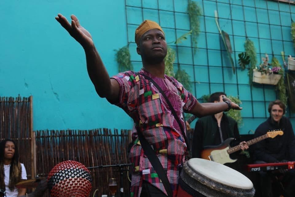 Cheick Bangoura stands in front of a blue wall. He holds a djumbe and opens his arms. He wears African styled shirt and hat. In the background there are three people and instruments.