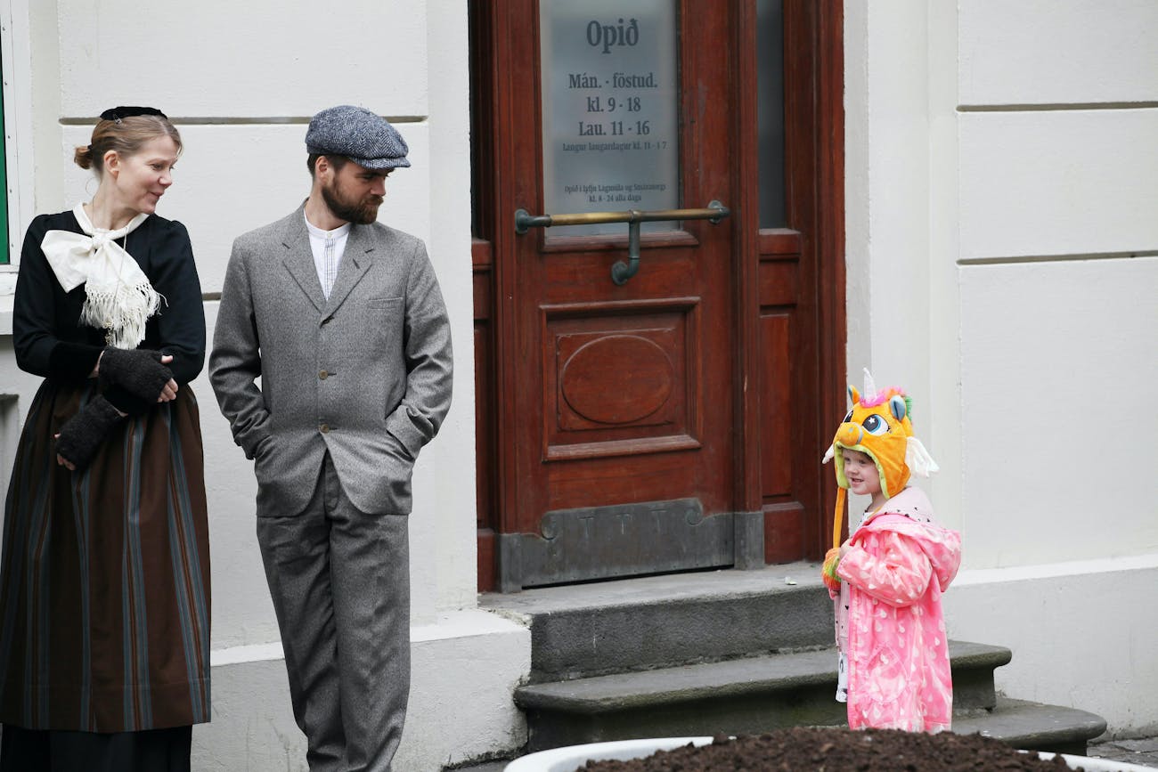 The photo was taken on Laugarvegurinn in front of an old pharmacy door. On the left side of the picture are a man and a woman dressed in old-fashioned clothes, sweaters and woolen jackets and trousers. They look at a child who is wearing a unicorn cap, gold and pink and wears a cap dress.