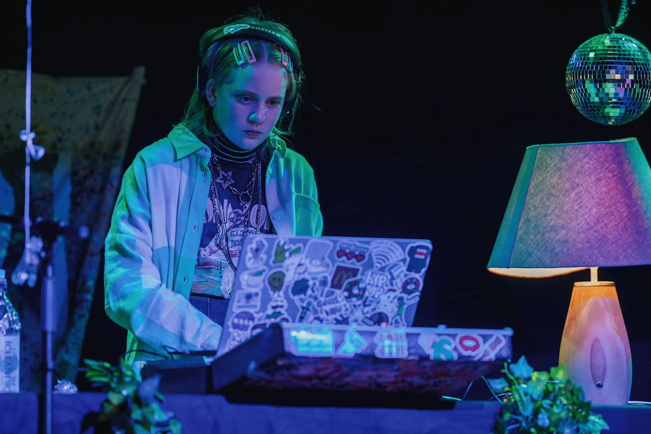 Picture of a young DJ looking in front of a computer screen with headphones on his head. The back of the computer is covered with stickers and the front has a mixer and a lamp. The image is illuminated with blue-green light.