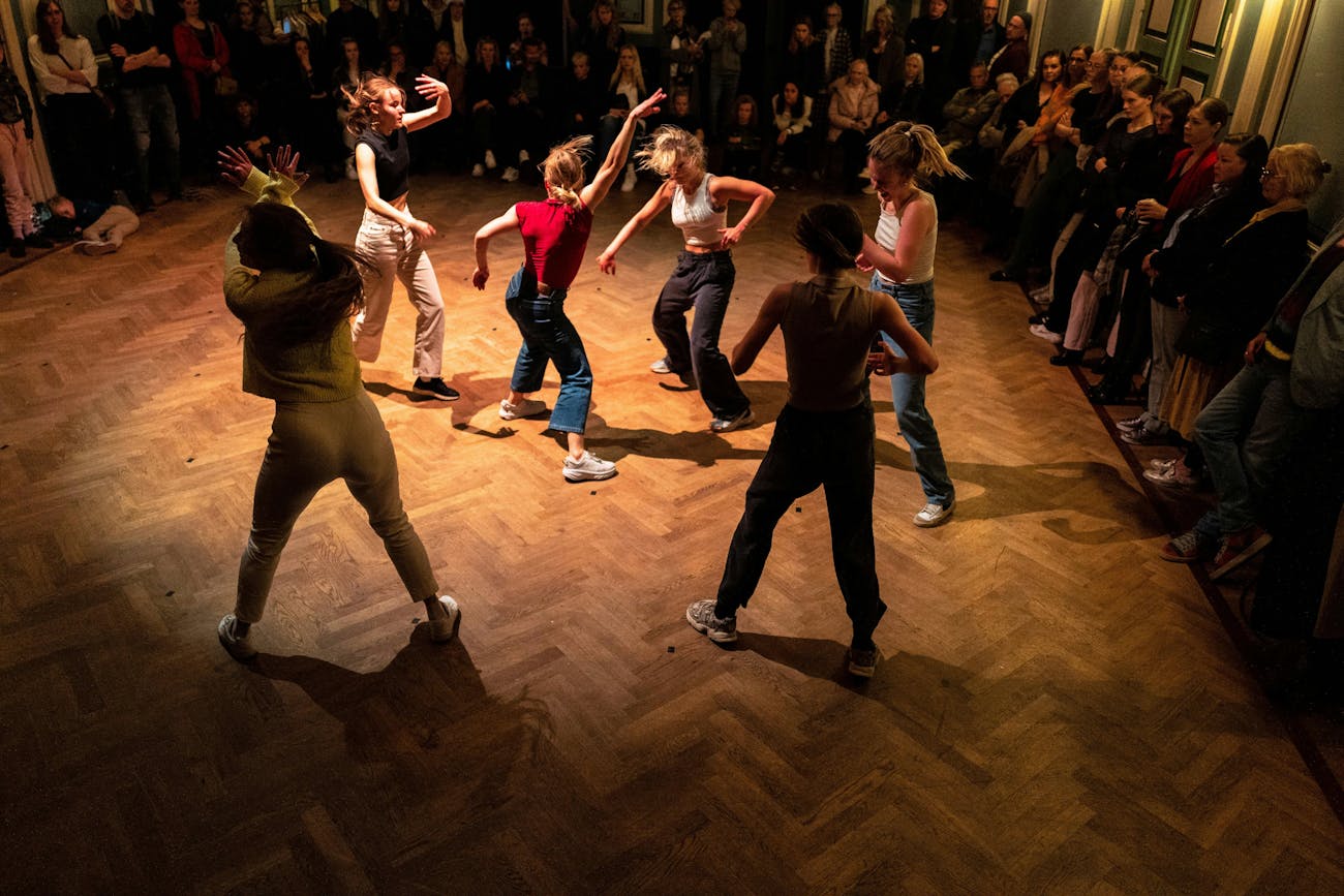 Six dancers in everyday clothes, dancing dynamically on a wooden floor. Audience members standing on the sides.