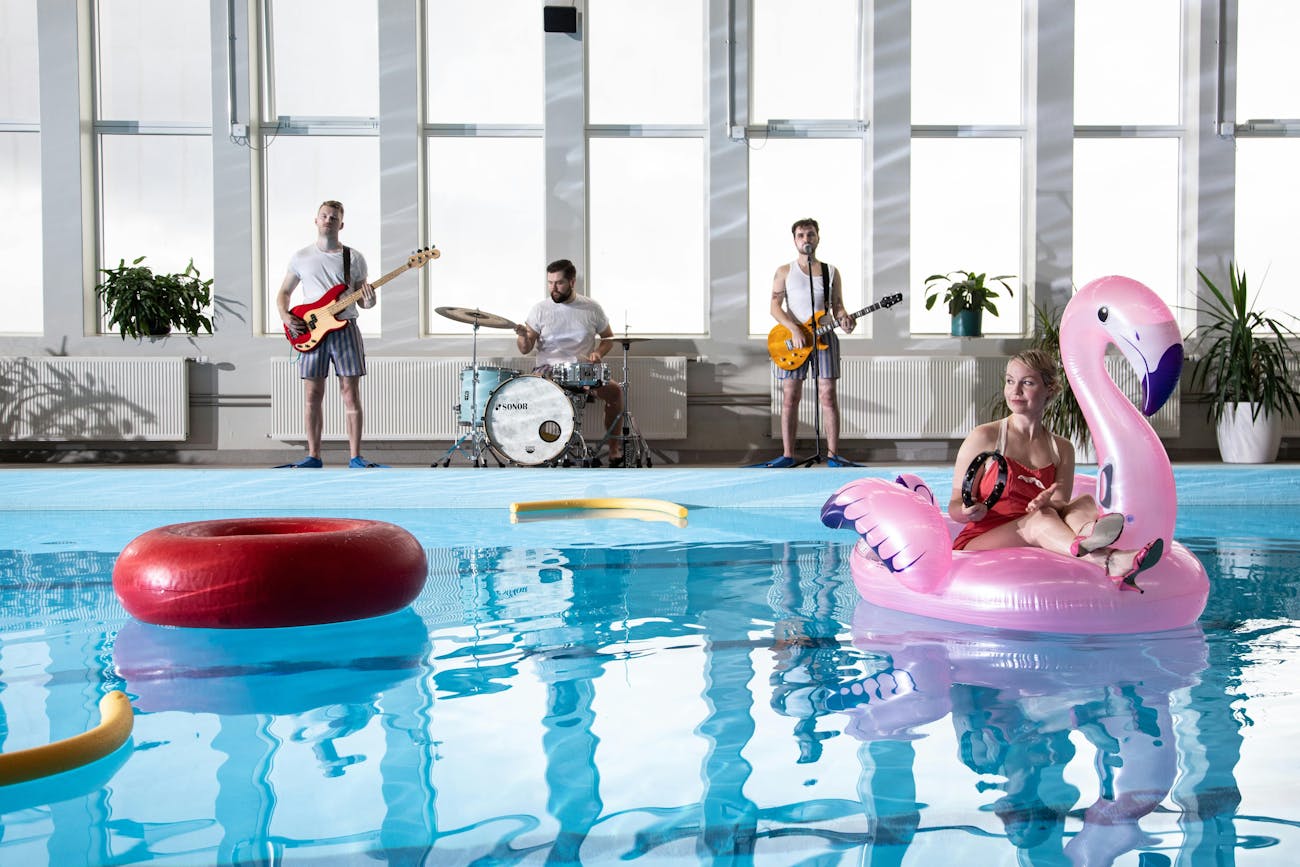There is a swimming pool in the foreground and windows in the background. In the background there are 3 people in white T-shirts and swimming trunks. They are. with guitar, bass and drum kit. There are plants on the windows and pool deck. In the pool there are yellow swimming noodles, one big red buoy and one inflated pink flamingo. On it sits a person in a red swimsuit and high heels holding a tambourine.