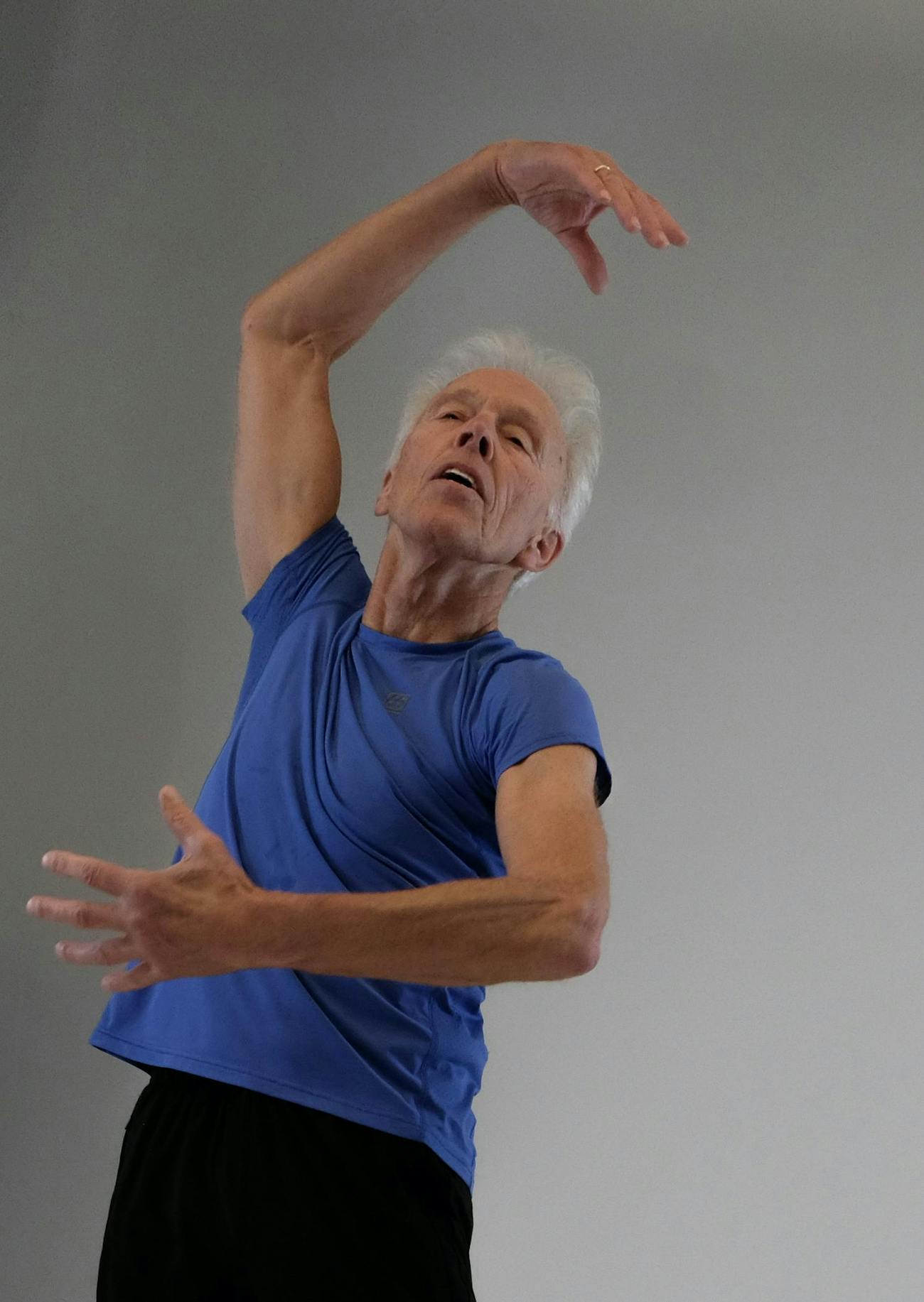 Image of a person dancing, one hand above their head and one hand in front of them. The person has gray hair. and closes his eyes, is wearing black pants and a blue top.