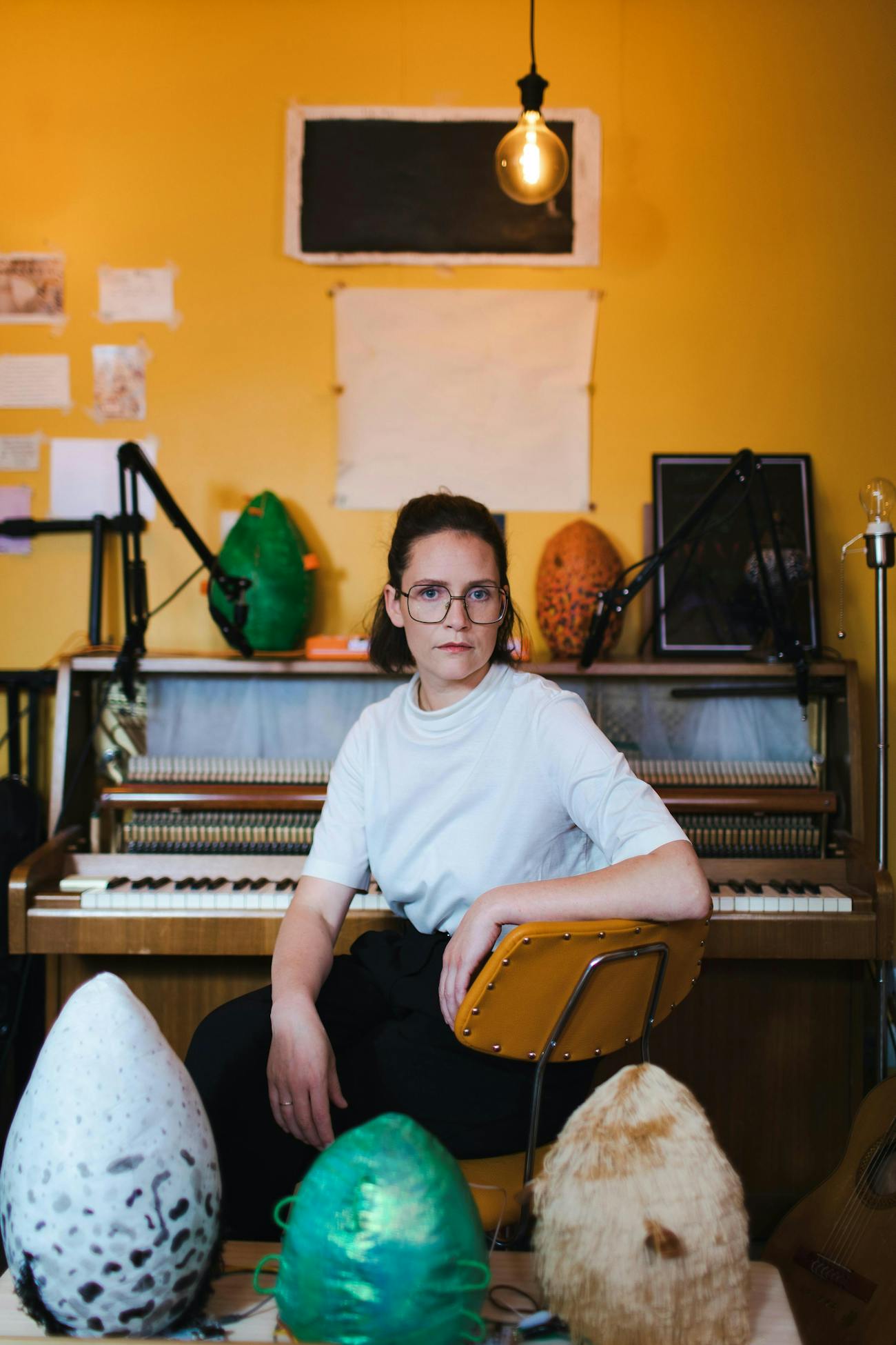 Sóley Stefánsdóttirsits in front of a piano. She looks at the camera. In the background there are two colorful sculptures resembling eggs.