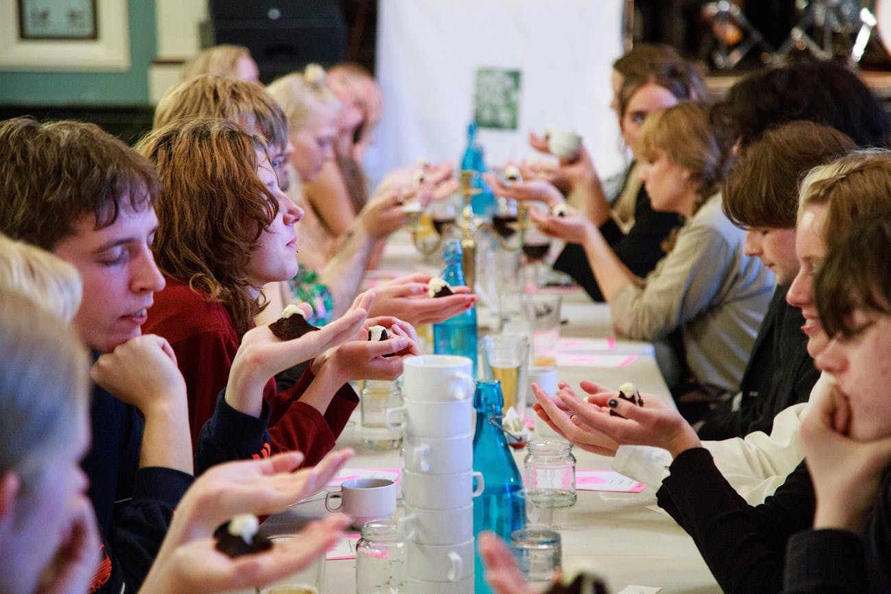 Guests sit at a long table. They hold cookies in open arms in front of them and close their eyes. On the table are blue bottles, glasses and cups.