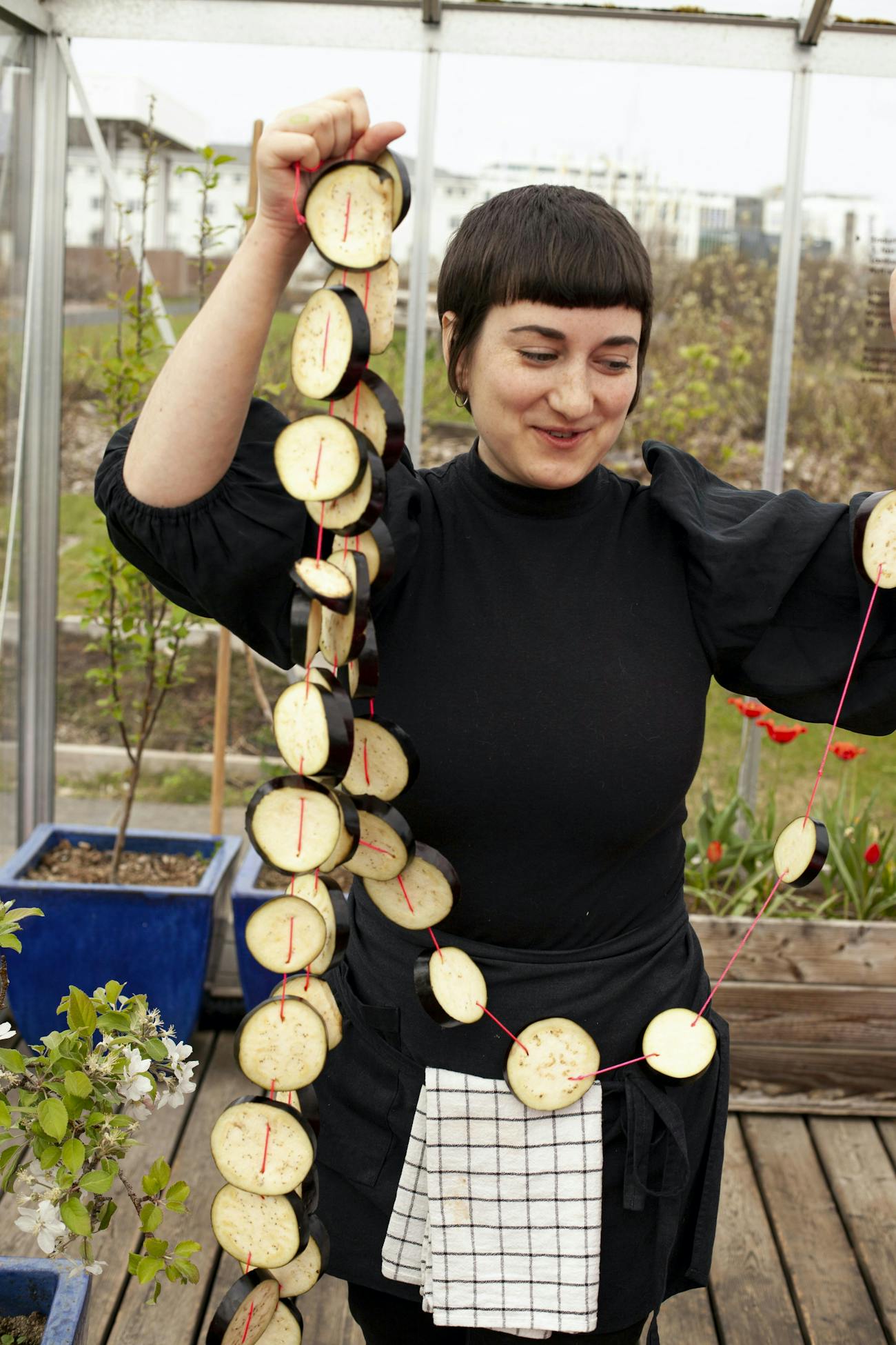 Pola Sutryk is in a greenhouse. Pola has short dark hair and wears a black dress and an apron with a kitchen towel hanging from it. Pola is holding a wreath made of eggplant slices and red thread. Pola smiles.