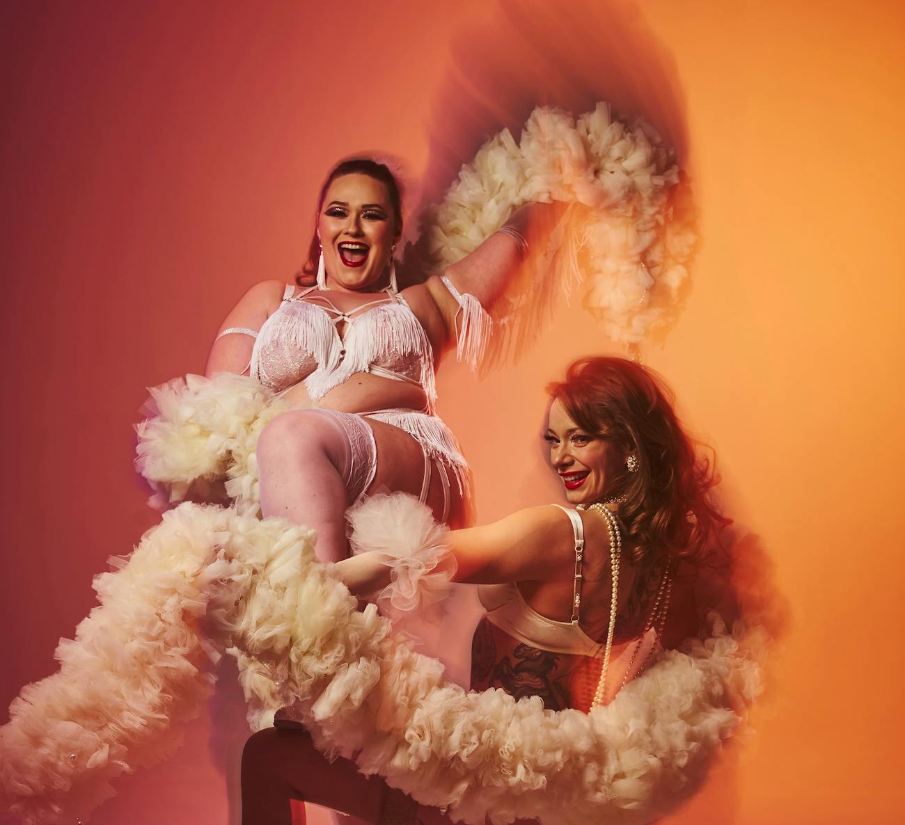 A pair of burlesque beauties pose before an orange background. They are both dark haired, light skinned and wearing white burlesque outfits with pearls, beads and fringe. They are both holding white boas which they wrap around themselves in a playful manner.